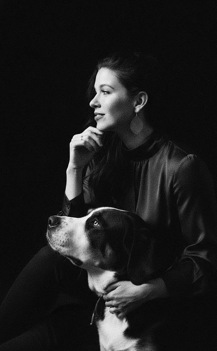 A black and white portrait of a woman with long hair, wearing earrings and a dark top, sitting with a dog that has black and white fur, while looking to the left.