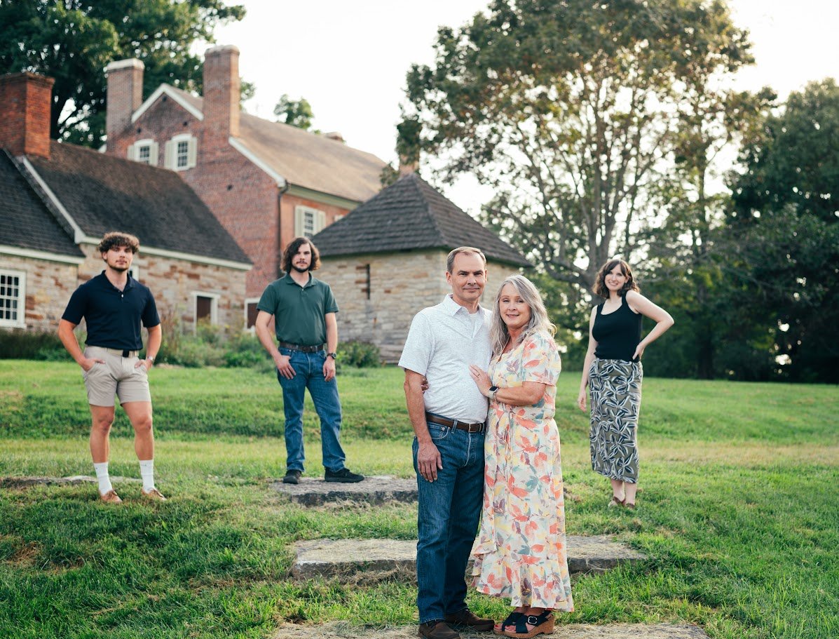 A family photo of five people standing on grass outside in front of historic brick and stone buildings with trees in the background. An older man and woman are in the front center, hugging. Three younger people stand behind them, spaced apart, with t