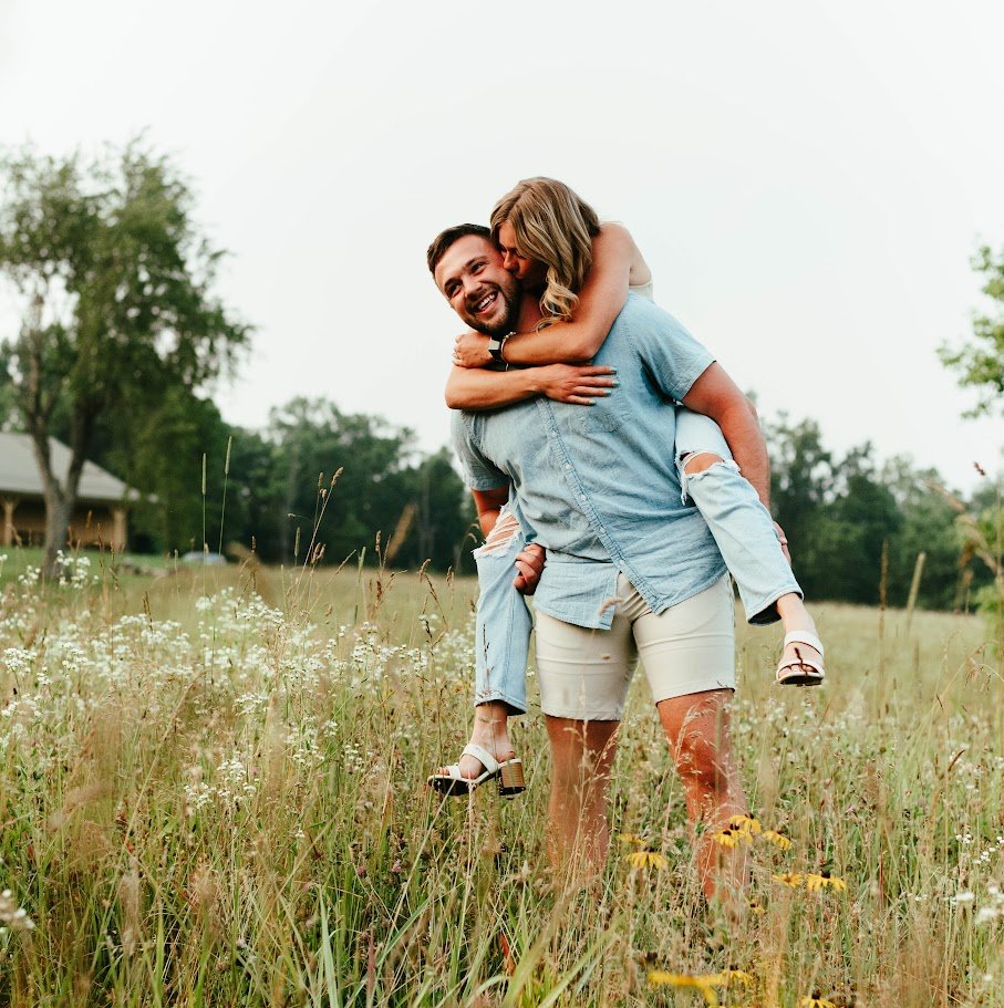 A man giving a piggyback ride to a woman in a field of grass and wildflowers, smiling and enjoying the outdoors.