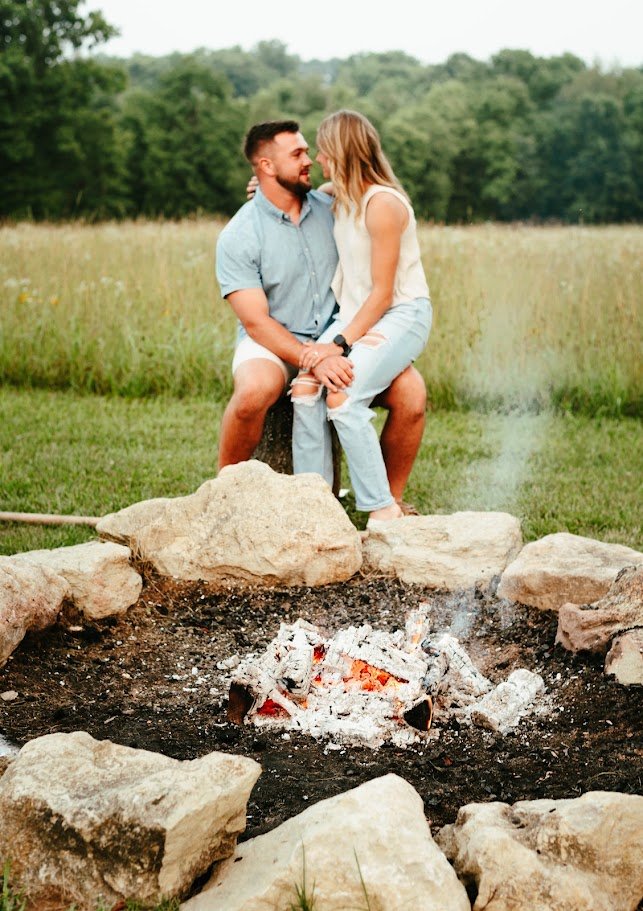 A couple sitting on a tree stump beside a campfire in a grassy field with trees in the background, standing close and gazing at each other.