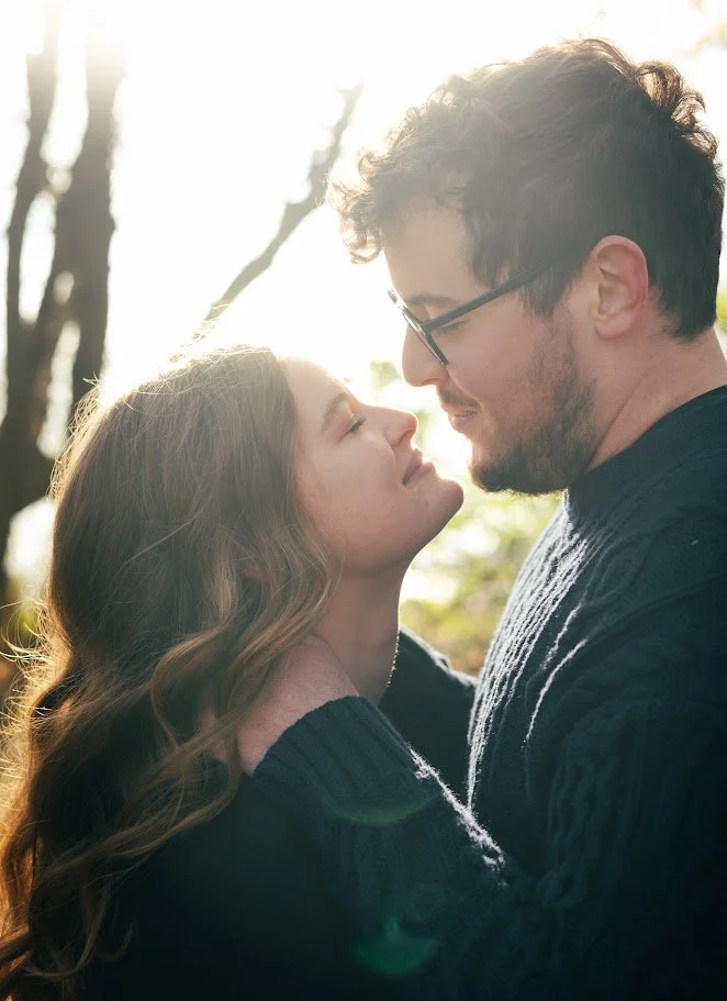 A couple gazing into each other's eyes outdoors with sunlight shining through trees in the background.