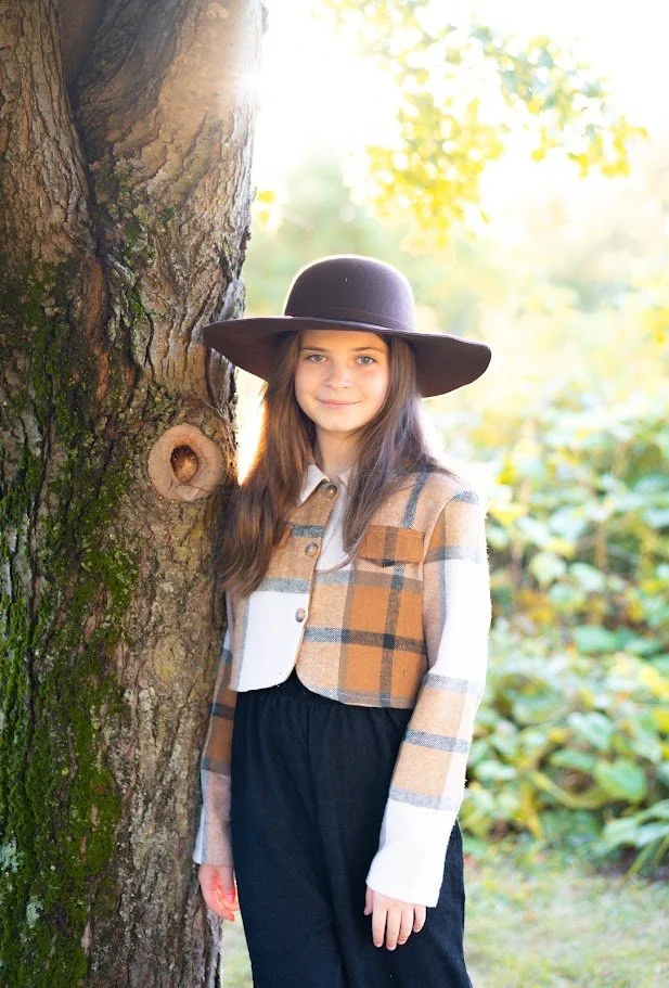 A young girl with long brown hair wearing a wide-brimmed hat, a plaid jacket, and dark pants, standing next to a tree with sunlight in the background.