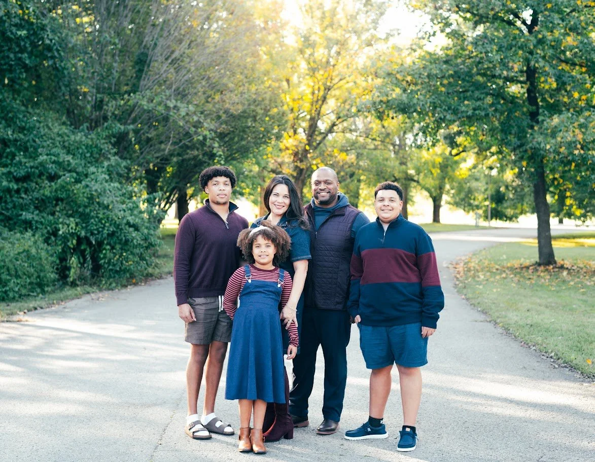 A family of five standing outdoors on a paved path surrounded by trees with fall foliage. The family includes a woman, a man, and three children, smiling and holding hands.