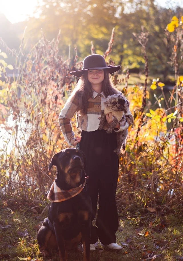 A girl with long brown hair wearing a large black hat, plaid shirt, and black pants standing outdoors by a body of water with autumn foliage in the background. She is holding a small puppy. A Rottweiler dog wearing a bandana sits in front of her.