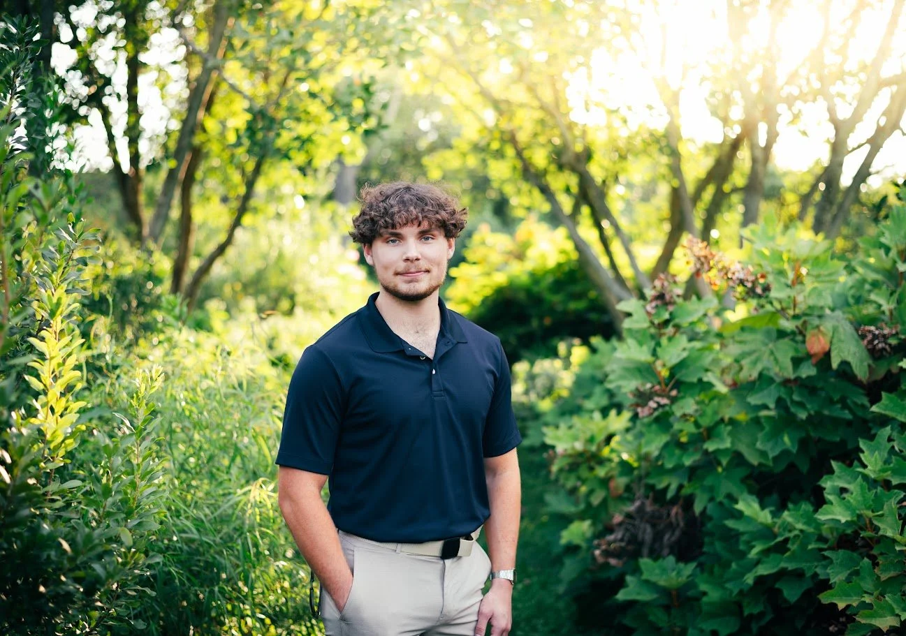 A young man in a navy blue polo shirt and beige pants standing in a lush green garden with sunlight filtering through the trees.