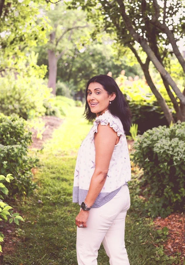 A woman with black hair smiling and looking at the camera, standing outdoors on a lush green pathway with trees and bushes around.