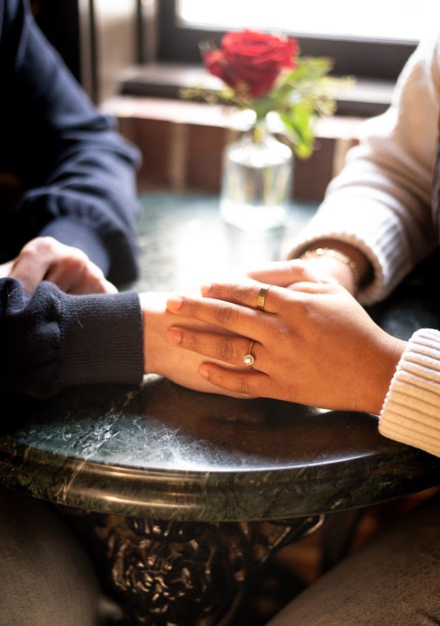 Two people holding hands on a dark marble table with a blurred window and flowers in the background.