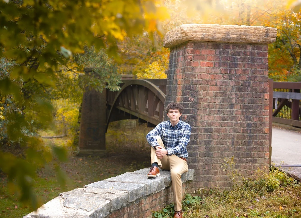 A young man in a plaid shirt and beige pants sitting on a stone wall, with a brick pillar and a wooden bridge in the background, surrounded by autumn trees.
