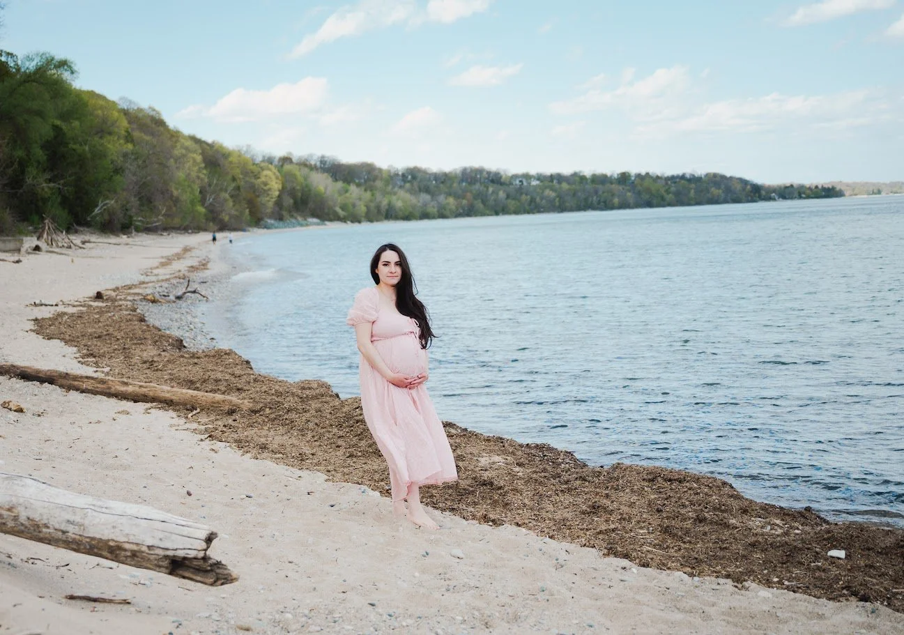Pregnant woman in a pink dress standing barefoot on a sandy beach by a body of water, with trees and a clear sky in the background.
