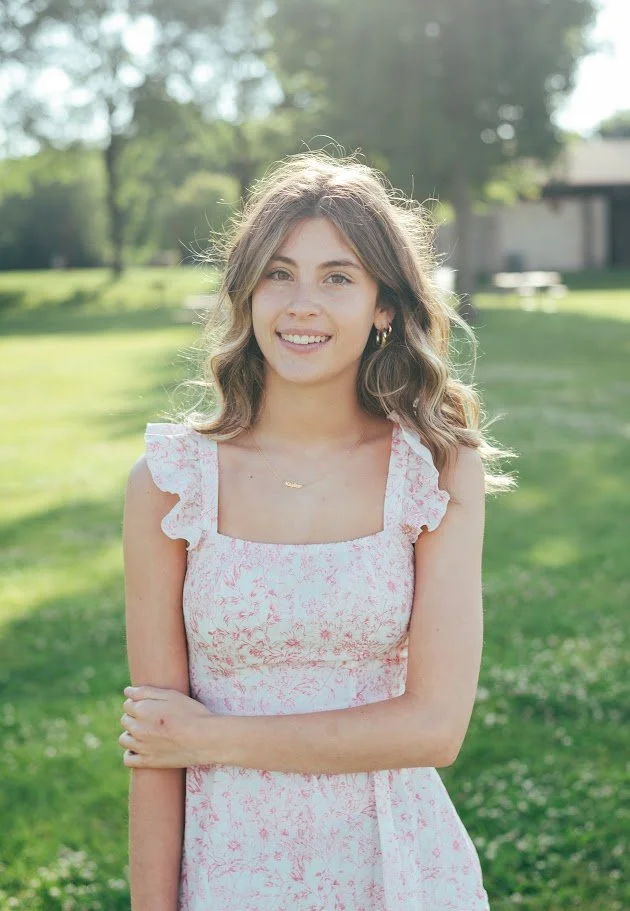 A young woman with wavy brown hair and hoop earrings standing outdoors on a grassy area with trees in the background, smiling at the camera.