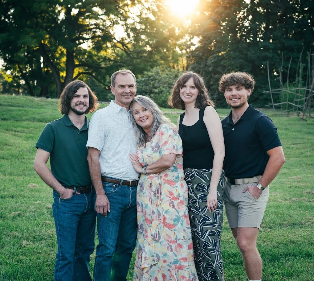 A family of five standing outdoors on a grassy field with trees and sunlight in the background, smiling at the camera.