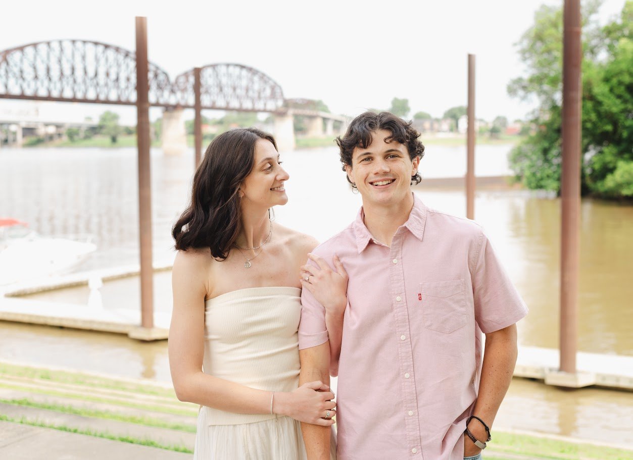 A smiling young man and a woman standing by a river with a bridge in the background. The woman is looking at the man and holding his arm, while the man is looking at the camera.