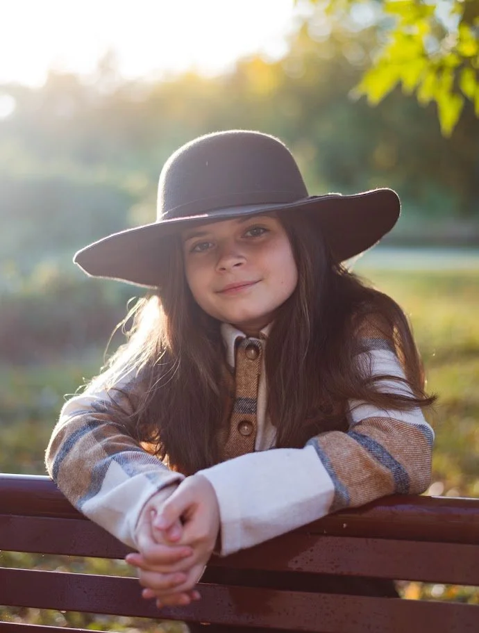 Young girl with long dark hair wearing a wide-brimmed hat and a plaid shirt, sitting on a park bench outdoors during sunset.