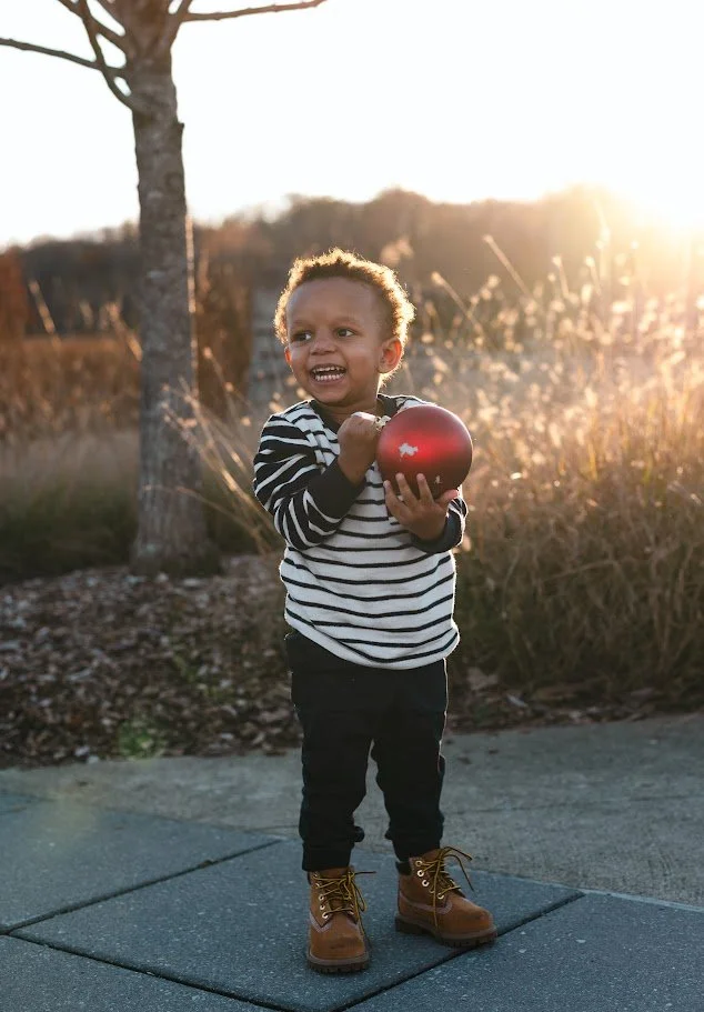 Young child smiling and holding a red Christmas ornament outdoors during sunset, wearing a striped shirt, black pants, and brown boots.