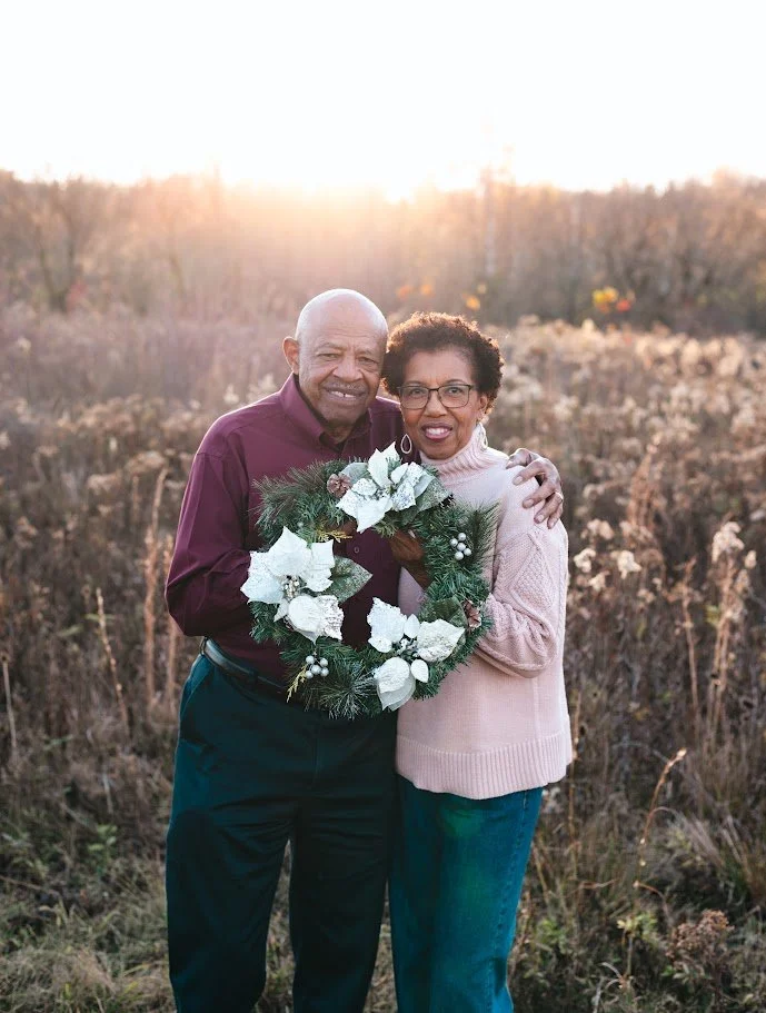 A happy elderly couple outdoors at sunset, holding a Christmas wreath decorated with white poinsettias and pine needles, standing in a field with dry grass.