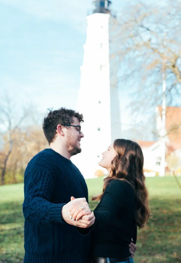 A young couple dancing outdoors near a lighthouse with trees and a grassy area in the background.