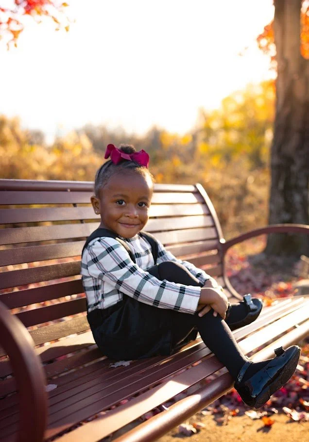 A young girl with a pink bow in her hair sitting on a park bench, smiling, surrounded by autumn trees and fallen leaves.