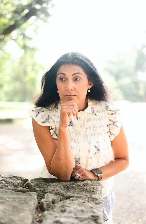A woman leaning on a stone surface outdoors, deep in thought, with trees and bright sunlight in the background.