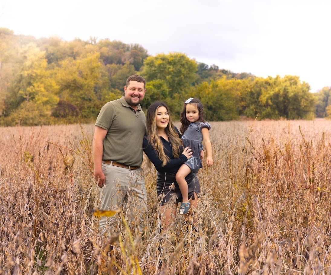 A family of three standing in a field of dried grass, with trees and a cloudy sky in the background.