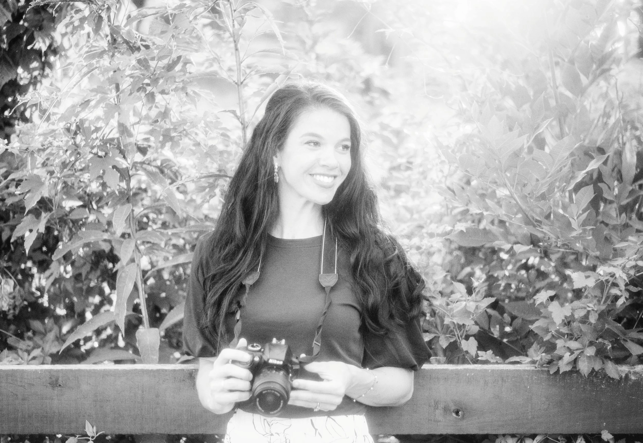 A woman with long dark hair smiling while holding a camera, standing in front of dense foliage.