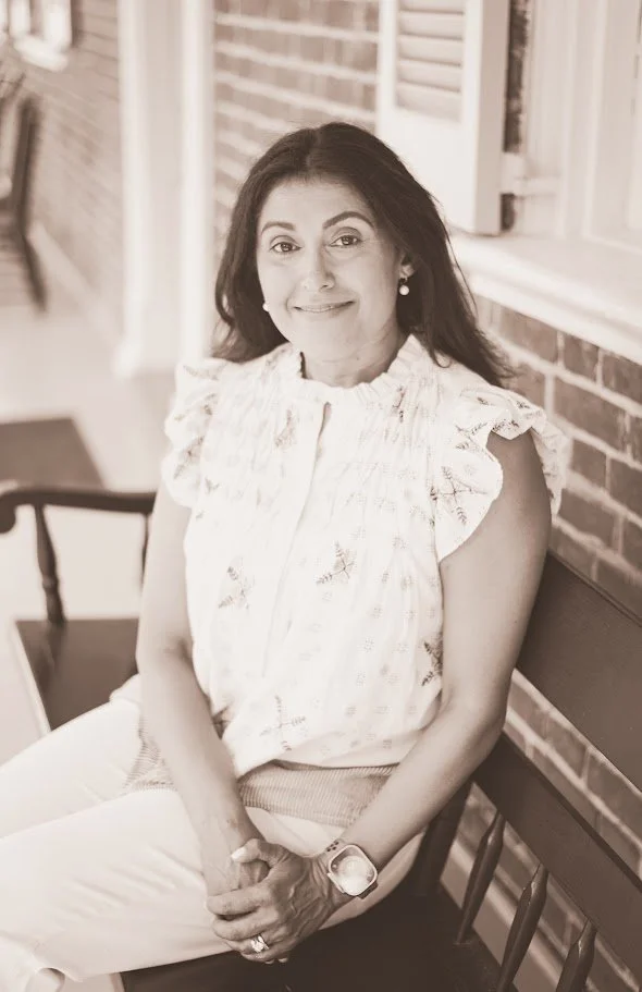 A woman with dark hair, smiling, wearing a light-colored, ruffled blouse and beige pants, sitting on a bench against a brick wall.