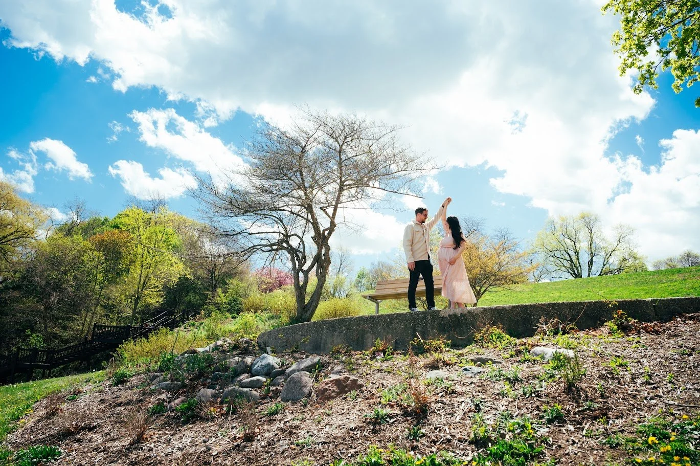 A couple standing on a grassy hill in a park, with the man twirling the woman under his arm. The sky is bright with scattered clouds, and trees with fresh spring leaves surround them.