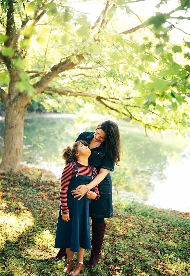 A woman and a young girl sharing a happy moment outdoors under a large tree by a lake, with sunlight filtering through the leaves.