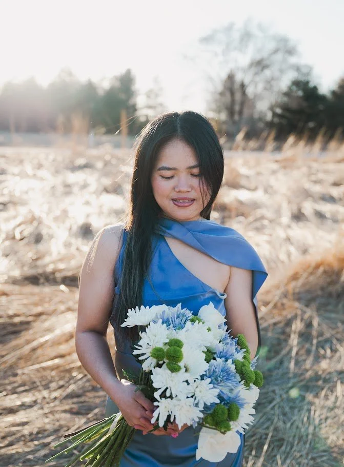 Woman in a blue dress holding a bouquet of white, blue, and green flowers outdoors during sunset.