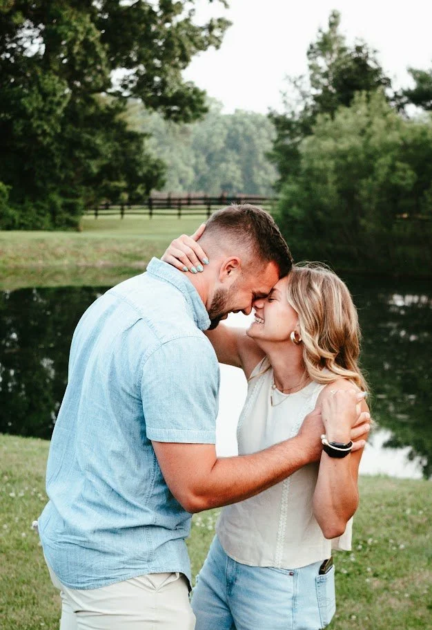A couple is outdoors near a river or pond, smiling and touching foreheads as they embrace. The man is wearing a light blue shirt and the woman is wearing a white sleeveless top and denim shorts. There are trees and a fence in the background.