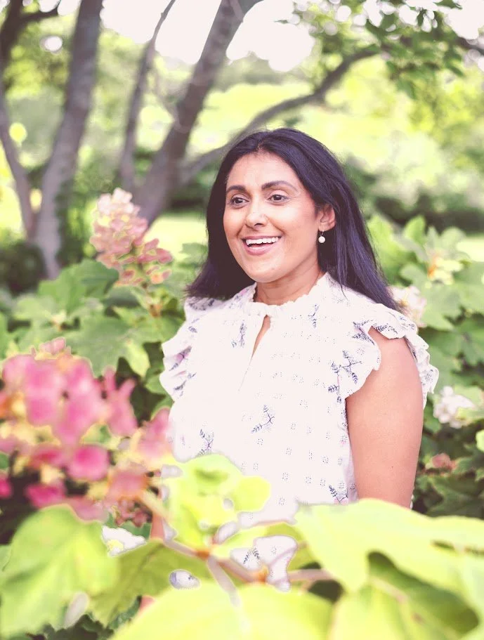 A woman with dark hair smiling outdoors among green foliage and pink flowers.