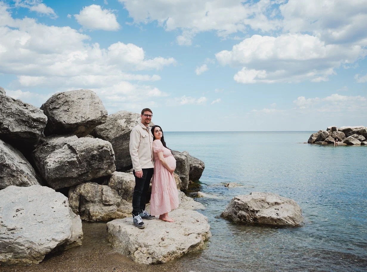 A man and a pregnant woman standing on rocks by the water at the beach, with clouds and blue sky in the background.