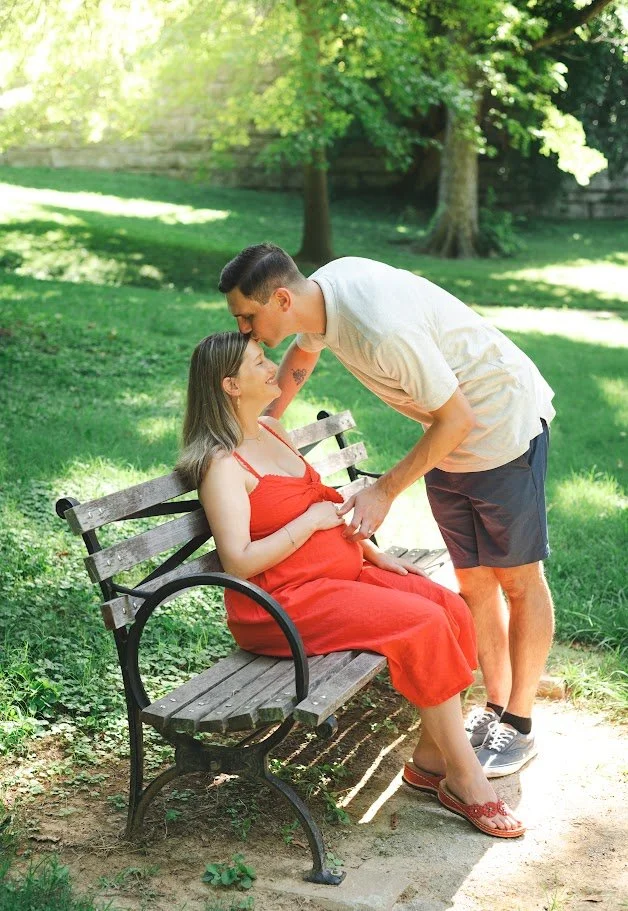 A couple sitting on a park bench, with the man leaning down to kiss the pregnant woman's forehead. The woman is wearing a red dress and slippers, and the man is dressed casually in a light-colored shirt and shorts. The scene is in a grassy, shaded pa
