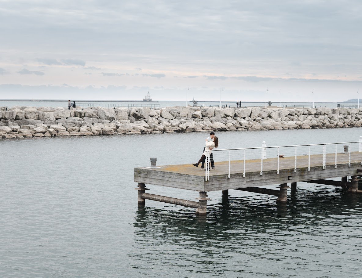 A couple embraces on a wooden dock by the water, with a rocky breakwater and lighthouse in the background, under a cloudy sky.