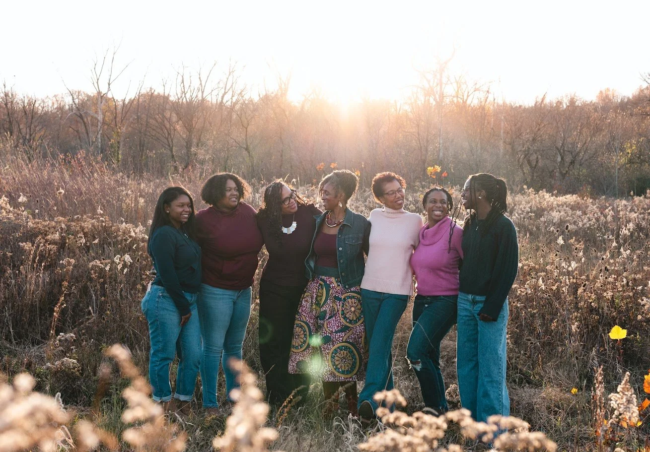 Seven women standing together in a field during sunset, smiling and enjoying each other's company.