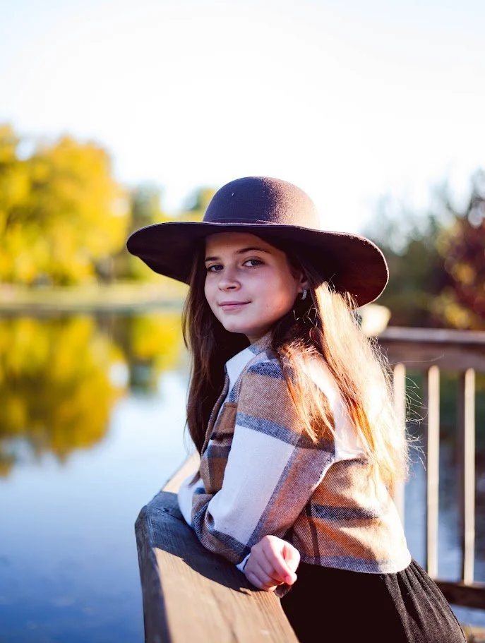 A young girl with long hair wearing a wide-brimmed hat and a plaid shirt, leaning on a wooden railing beside a calm lake during sunset with trees in the background.