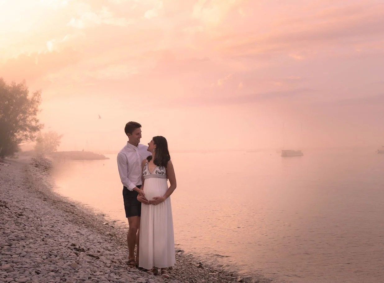 A couple standing on a rocky beach during sunset, with the woman pregnant, looking at each other, with a calm water body and a cloudy pink sky in the background.