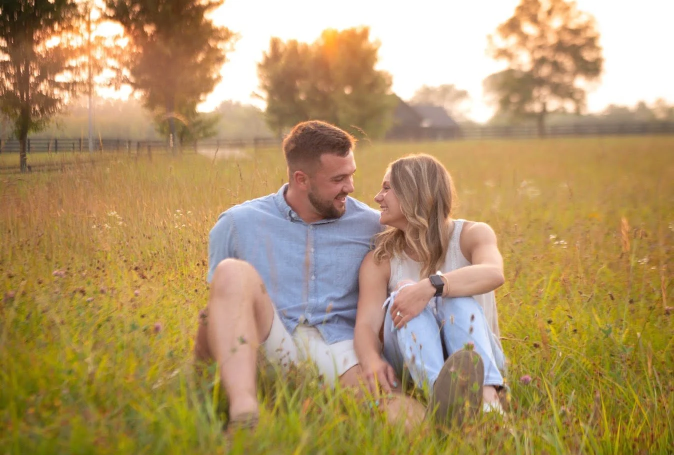 A young couple sitting together on a grassy field during sunset, looking at each other and smiling. The man is wearing a blue button-down shirt, and the woman is in a white top and light blue jeans.