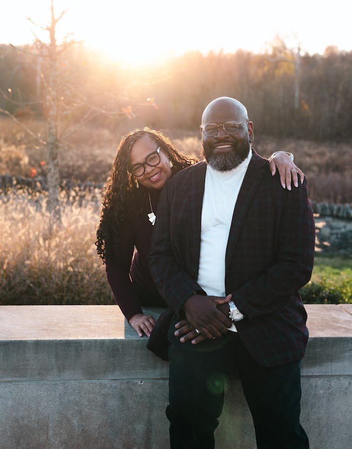 A man and woman smiling outdoors at sunset, with the woman resting her head on the man's shoulder, standing behind a concrete railing in a natural setting with trees and dry grass in the background.