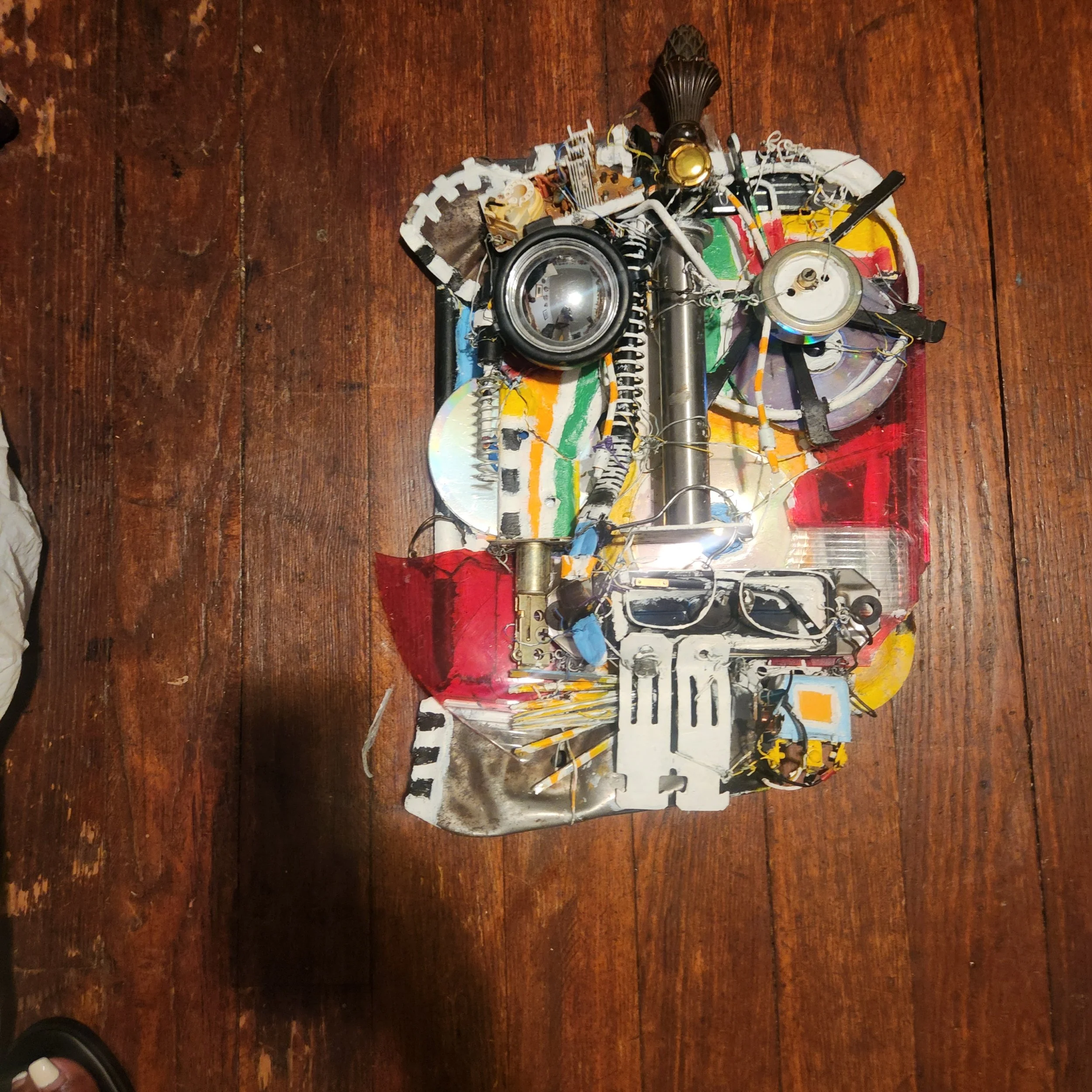 Found object artwork by Karl A. McIntosh featuring salvaged electronic components, wires, metal parts, and painted elements mounted on a wooden panel.
