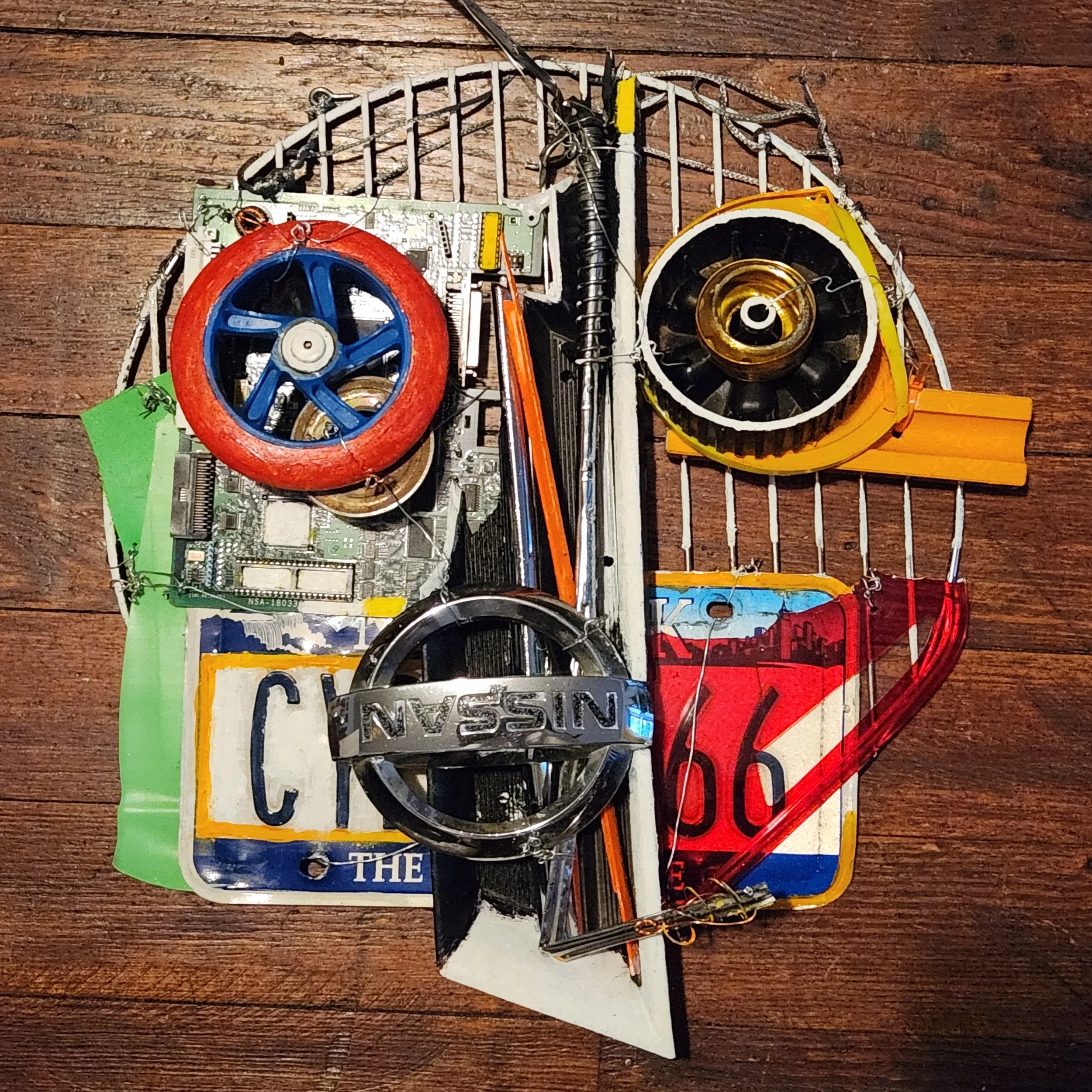 Found object artwork by Karl A. McIntosh featuring vintage license plates, salvaged electronic components, metal parts, and colorful recycled materials arranged on a wooden panel.