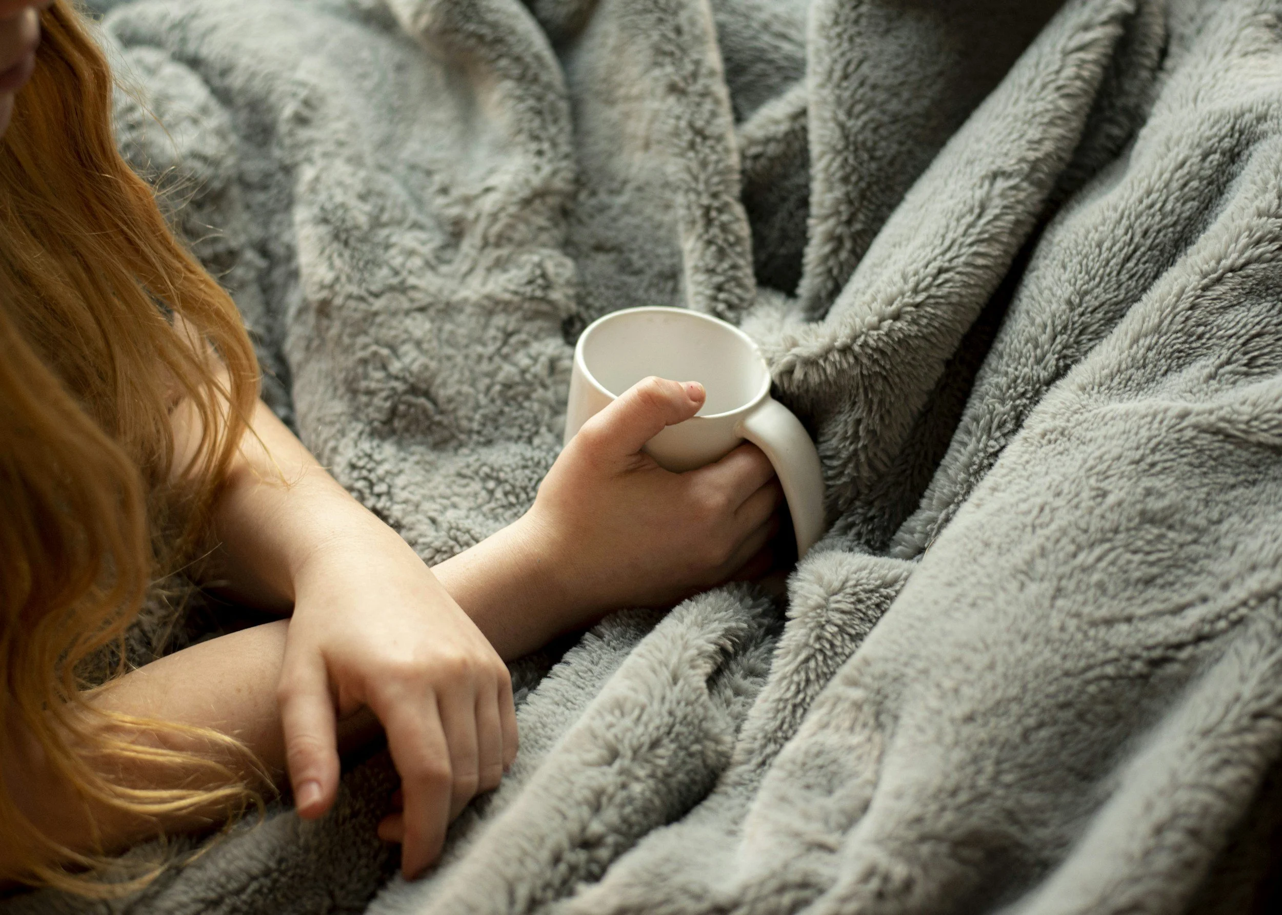 A person with red hair holding a white mug while lying on a cozy gray fleece blanket.