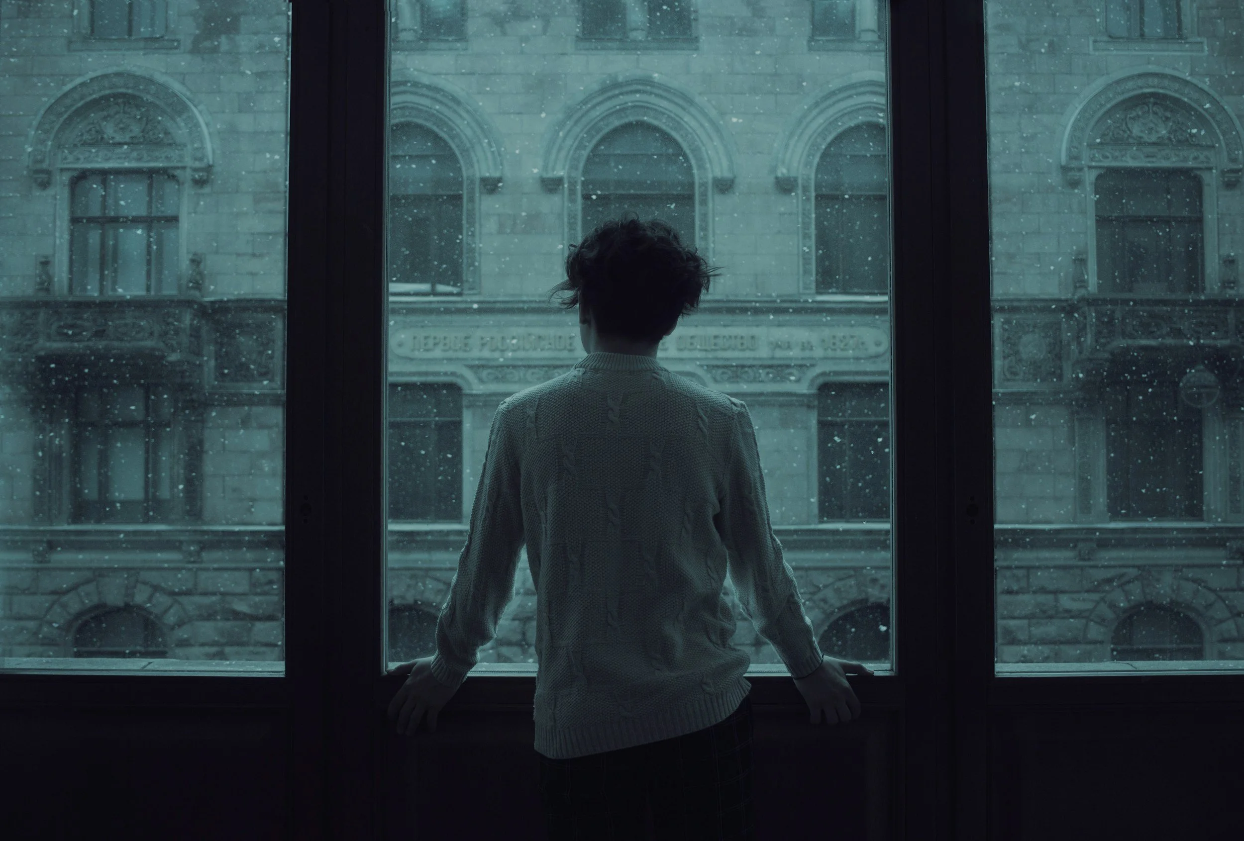 Person standing by a large window during a rainy day, looking outside at a historic building with arched windows.