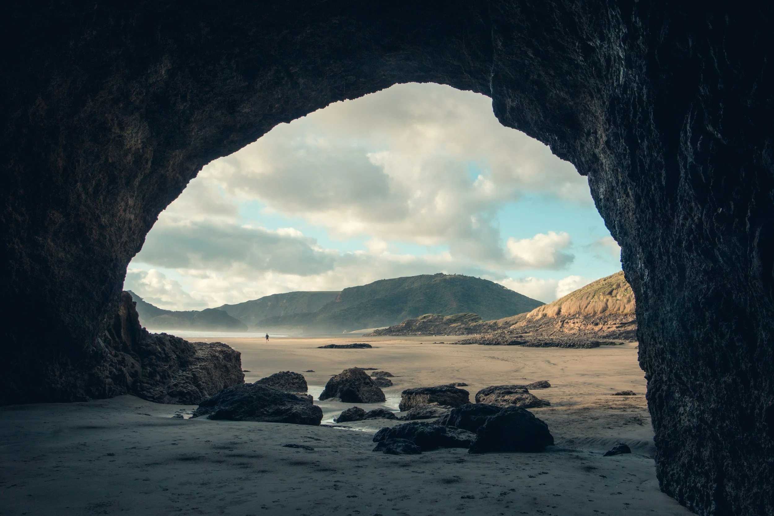 View through a large coastal cave showing a sandy beach, rocks, hills in the distance, cloudy sky, and a person walking on the beach.