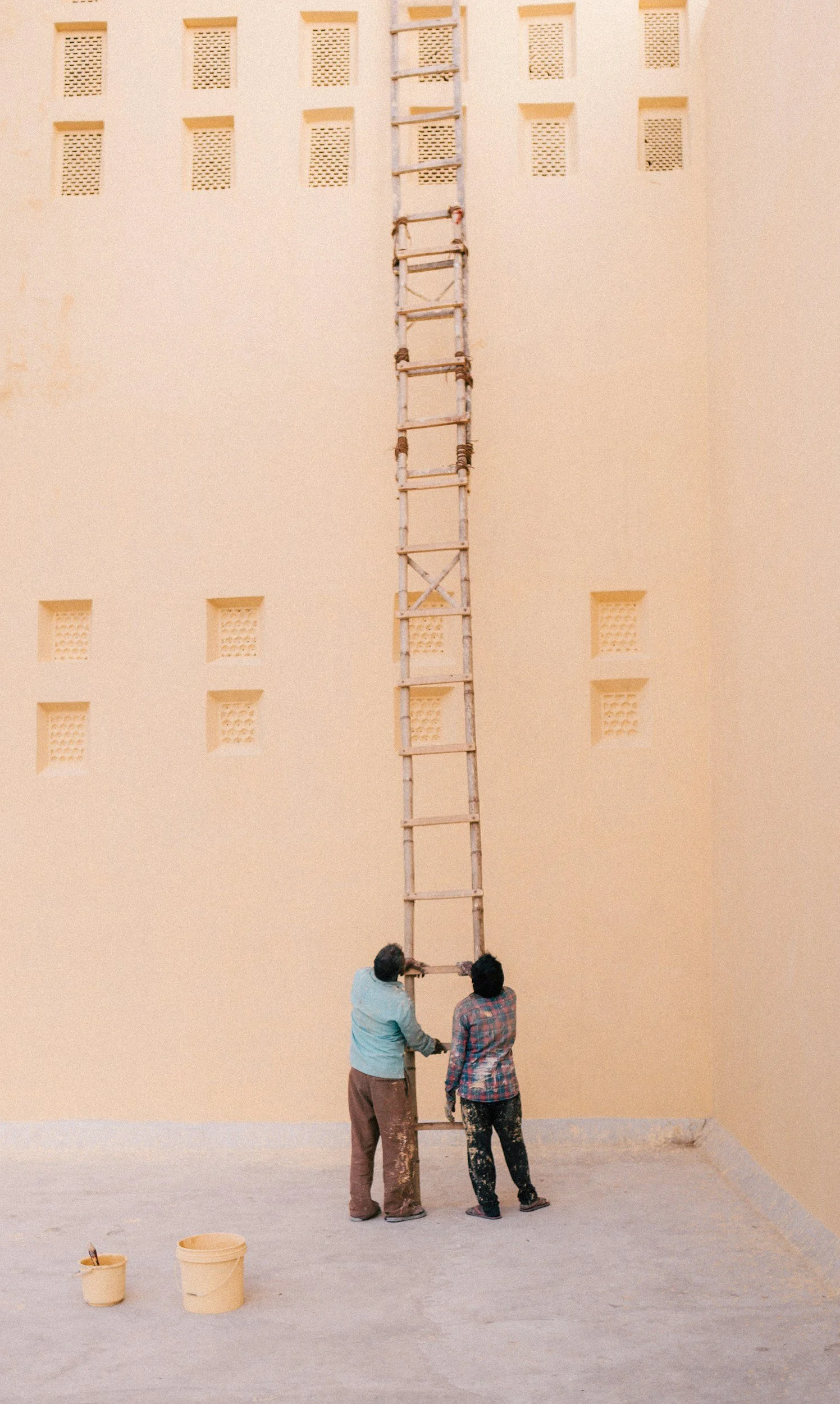 Two men are standing and holding a tall ladder against a beige wall with small square vents. There are two yellow buckets on the ground in front of them.