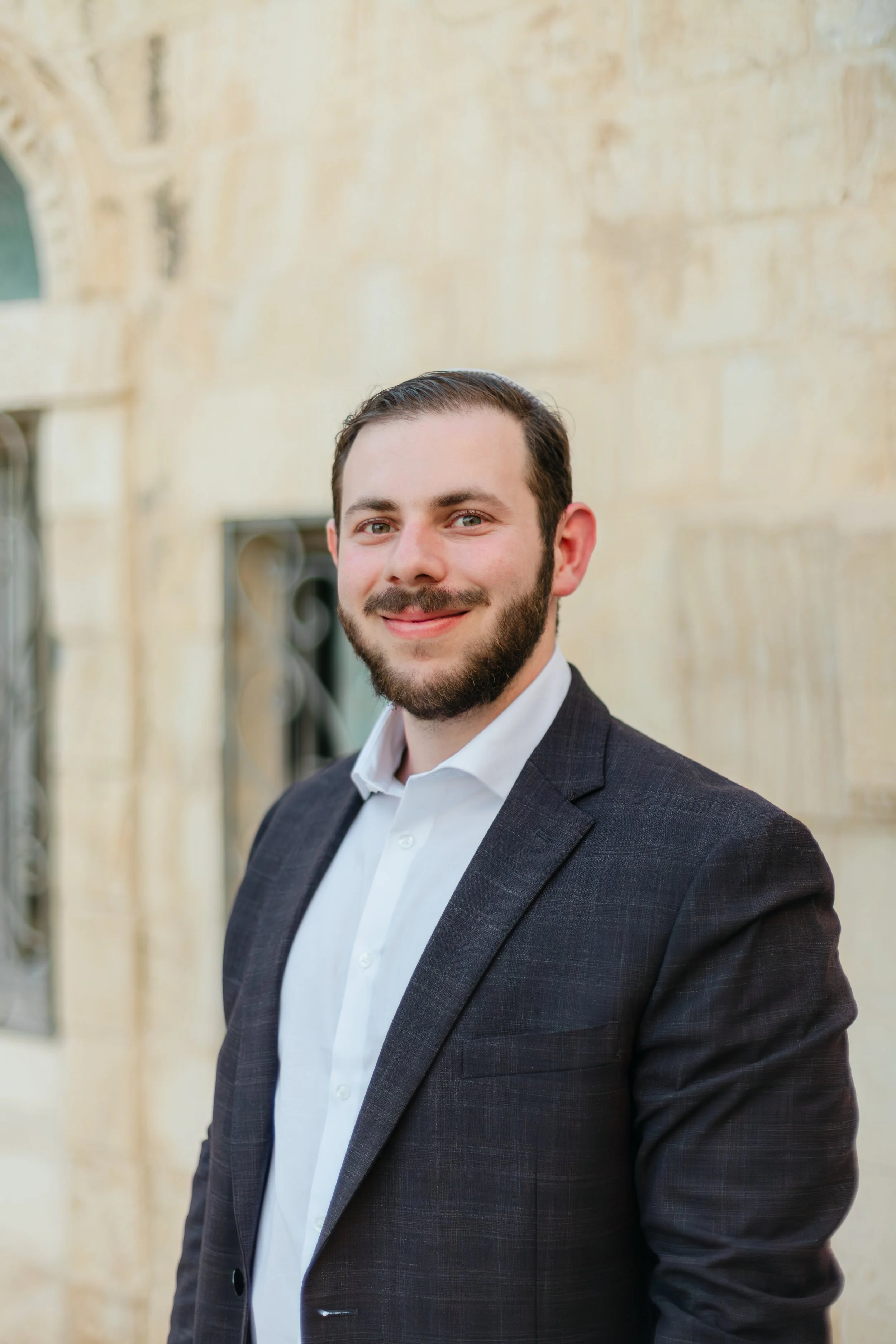 A therapist with dark hair, beard, and light skin wearing a white shirt and a dark suit jacket, smiling, standing outdoors against a beige stone or concrete wall with windows.