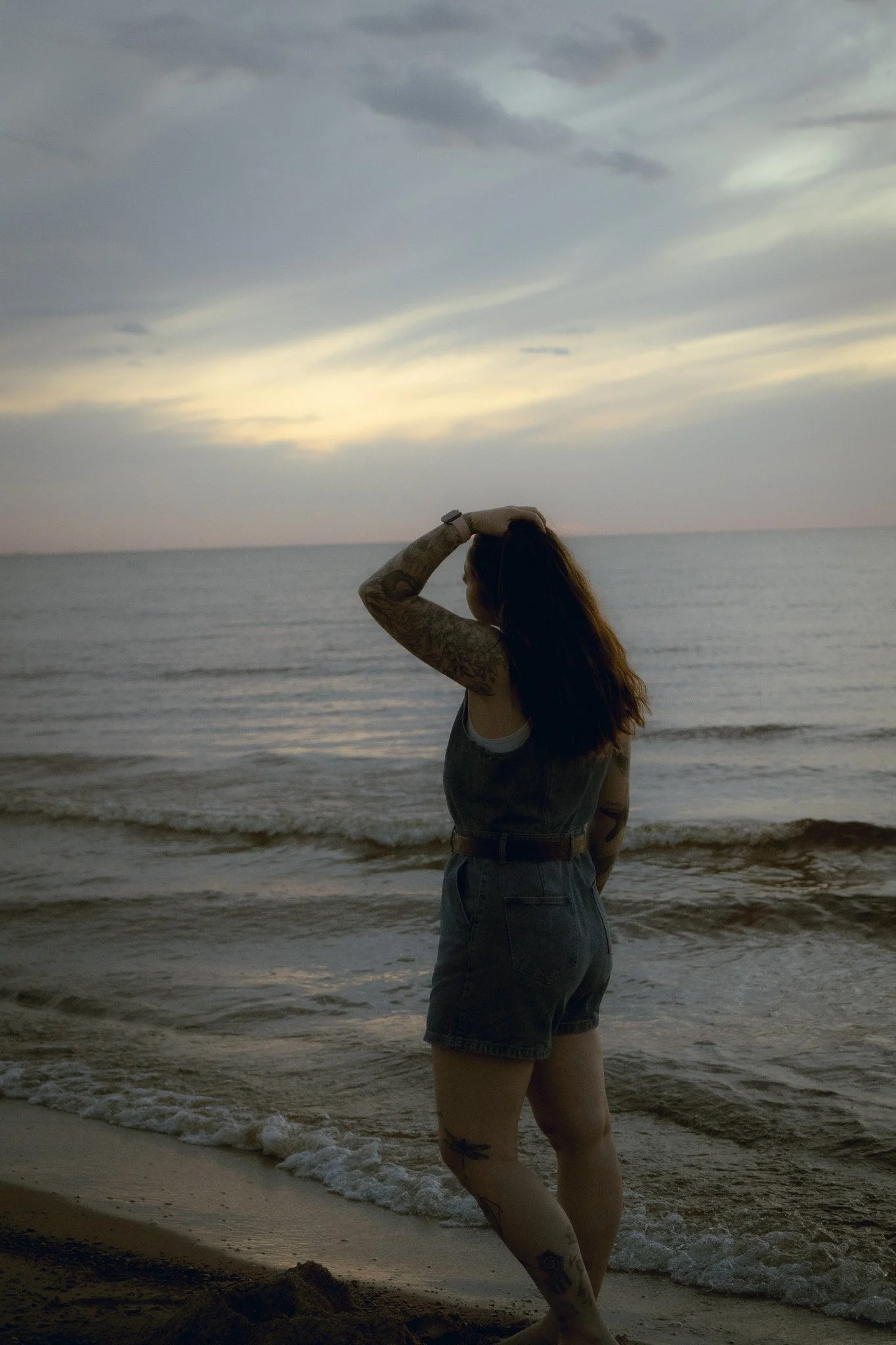 A woman with long dark hair and tattoos on her arm and leg standing on the beach near the water, looking towards the horizon during sunset or dusk, with clouds in the sky.