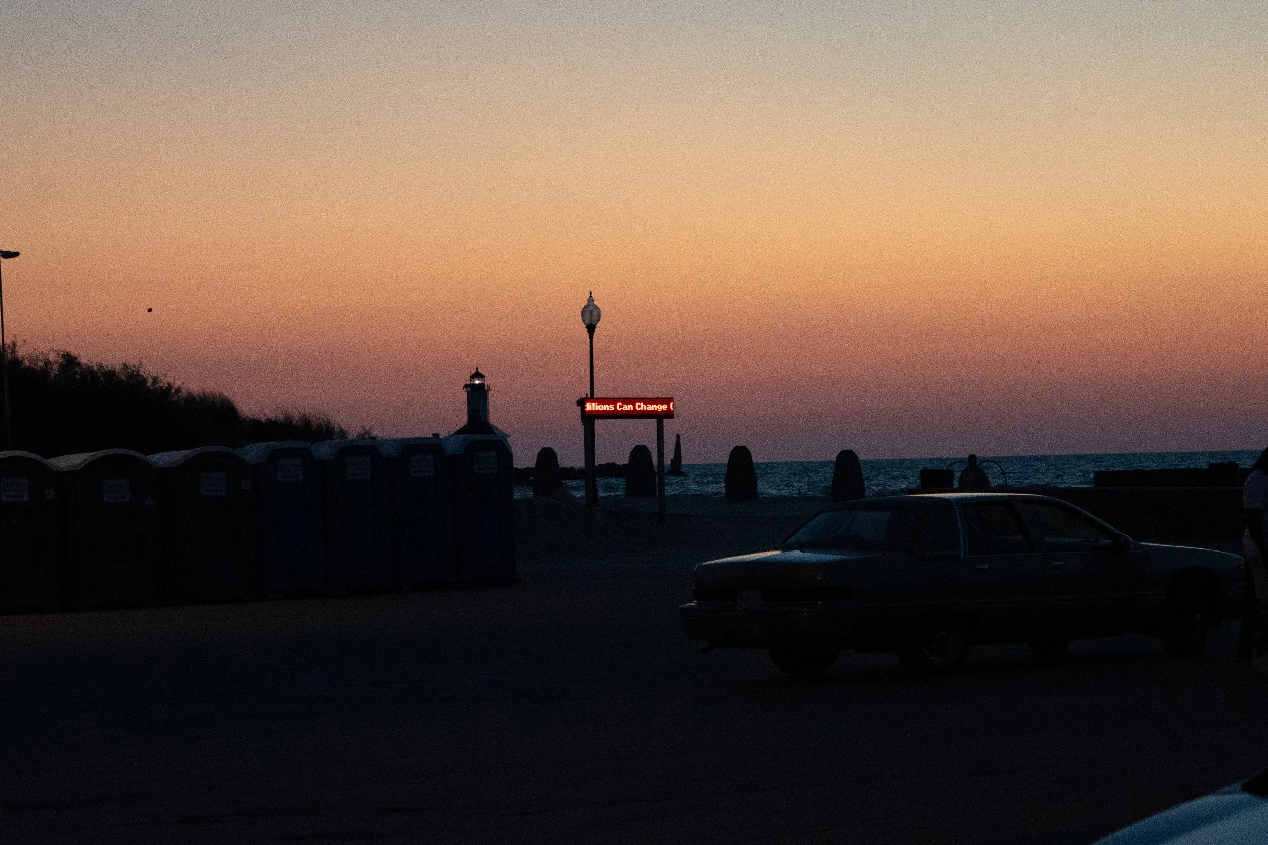 A sunset at the beach with a lighthouse, row of port-a-potties, parked car, lamppost, and illuminated sign that reads 'Situations Can Change'.