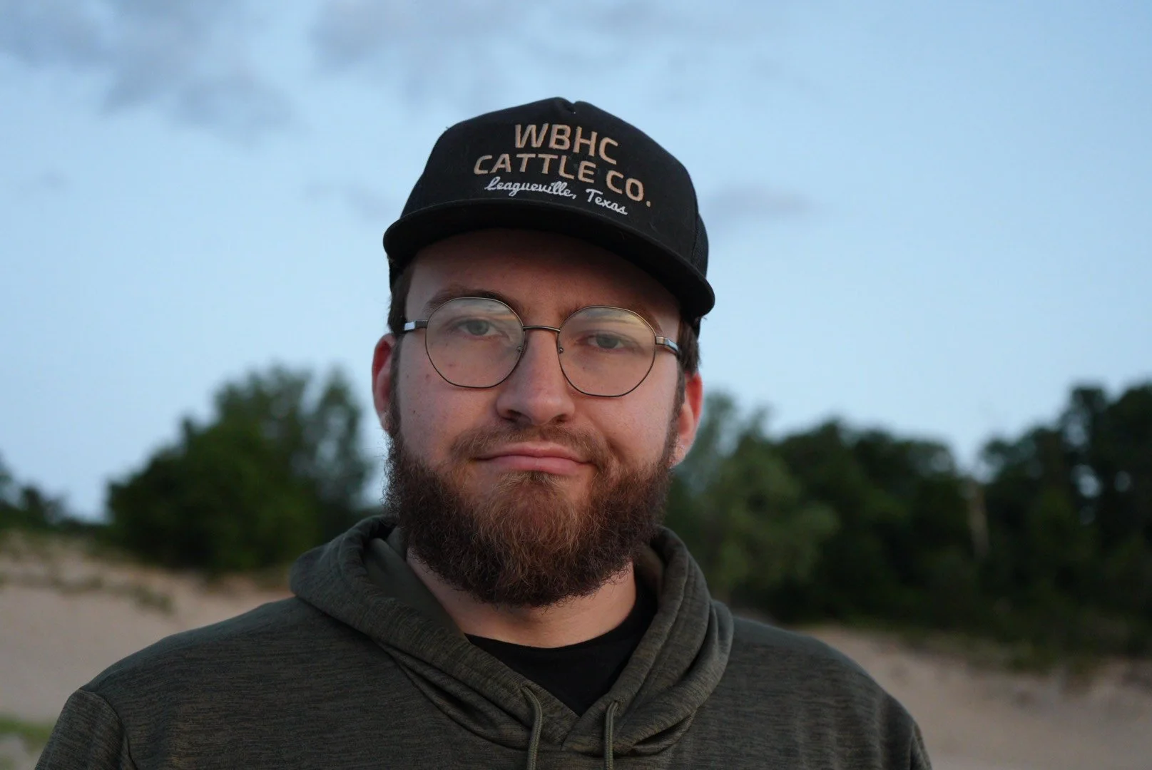 A man with glasses and a beard wearing a black baseball cap and a hoodie, standing outdoors with trees and a cloudy sky in the background.