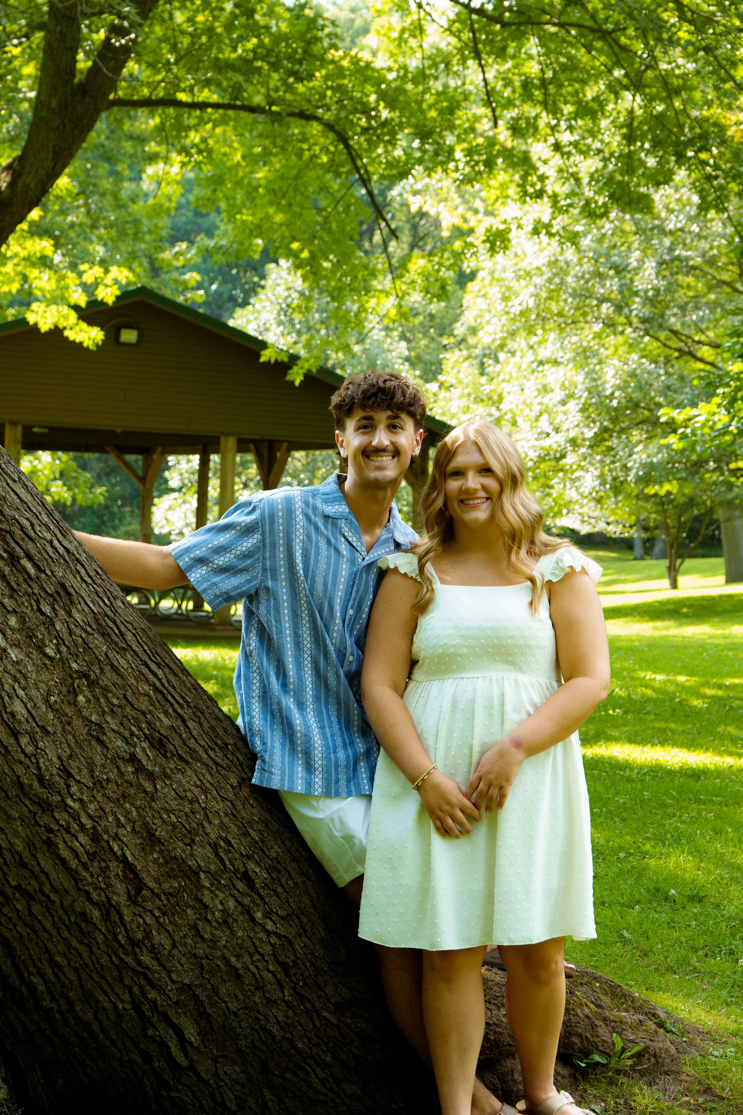 Two young adults, a man and a woman, standing outdoors in a park with lush green trees and grass, smiling at the camera. The man is wearing a blue striped shirt and white shorts, leaning against a large tree. The woman is in a light yellow dress, sta