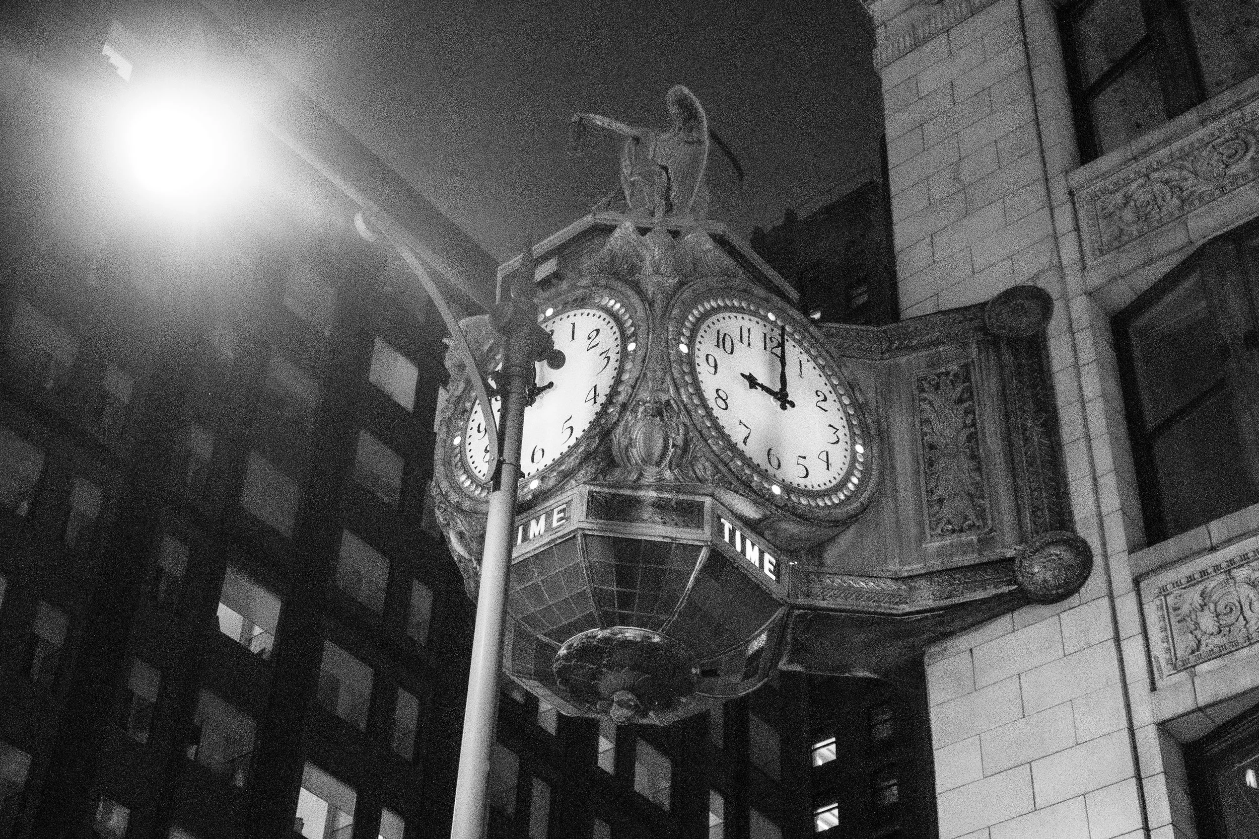 A vintage clock with two faces showing 1:42 at night, mounted on a brick building at night, illuminated by a bright streetlamp.